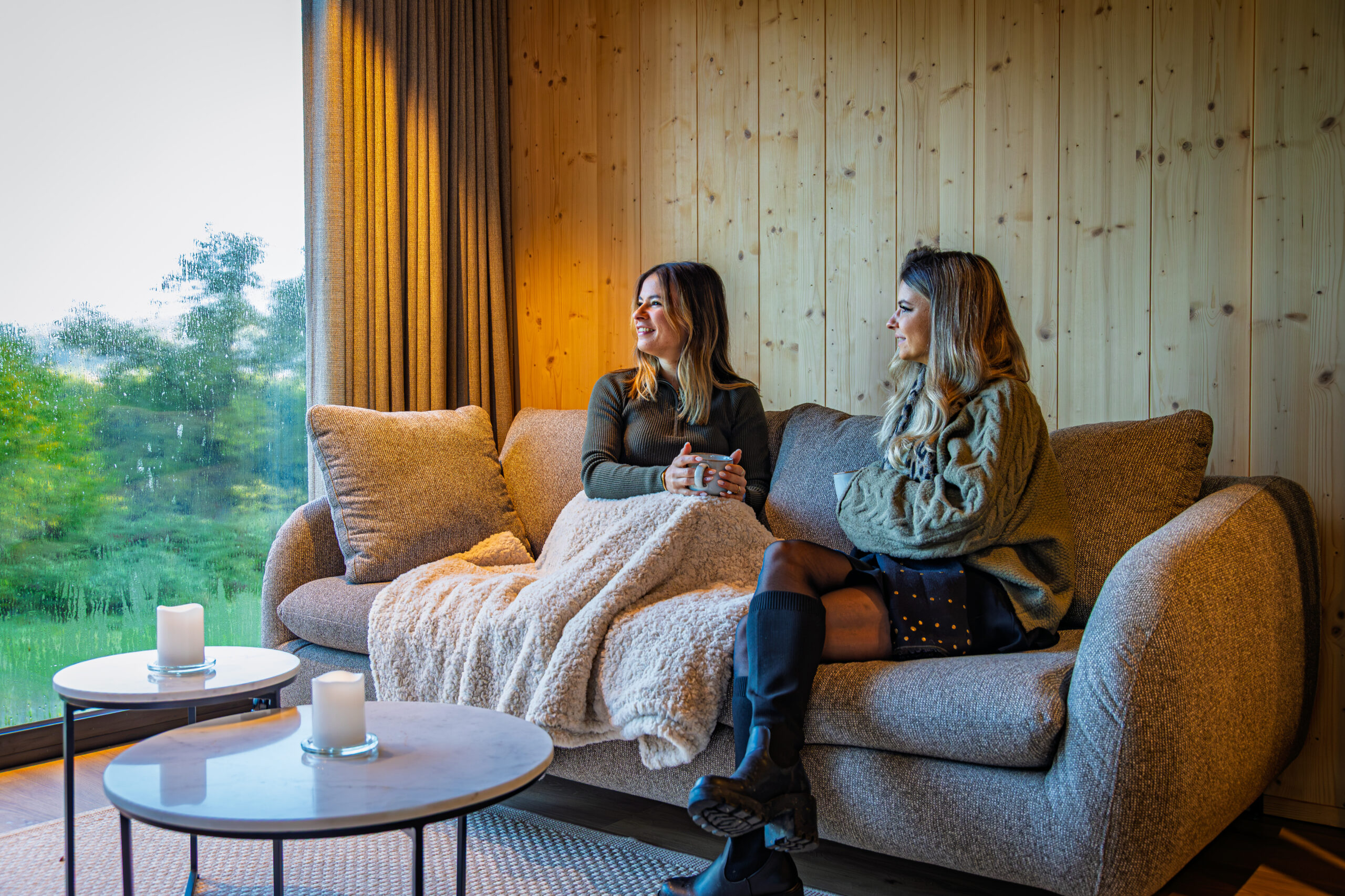 Deux femmes se détendent sous un plaid sur le canapé d'un lodge Les Hauts du Val, bougies allumées et vue sur la verdure