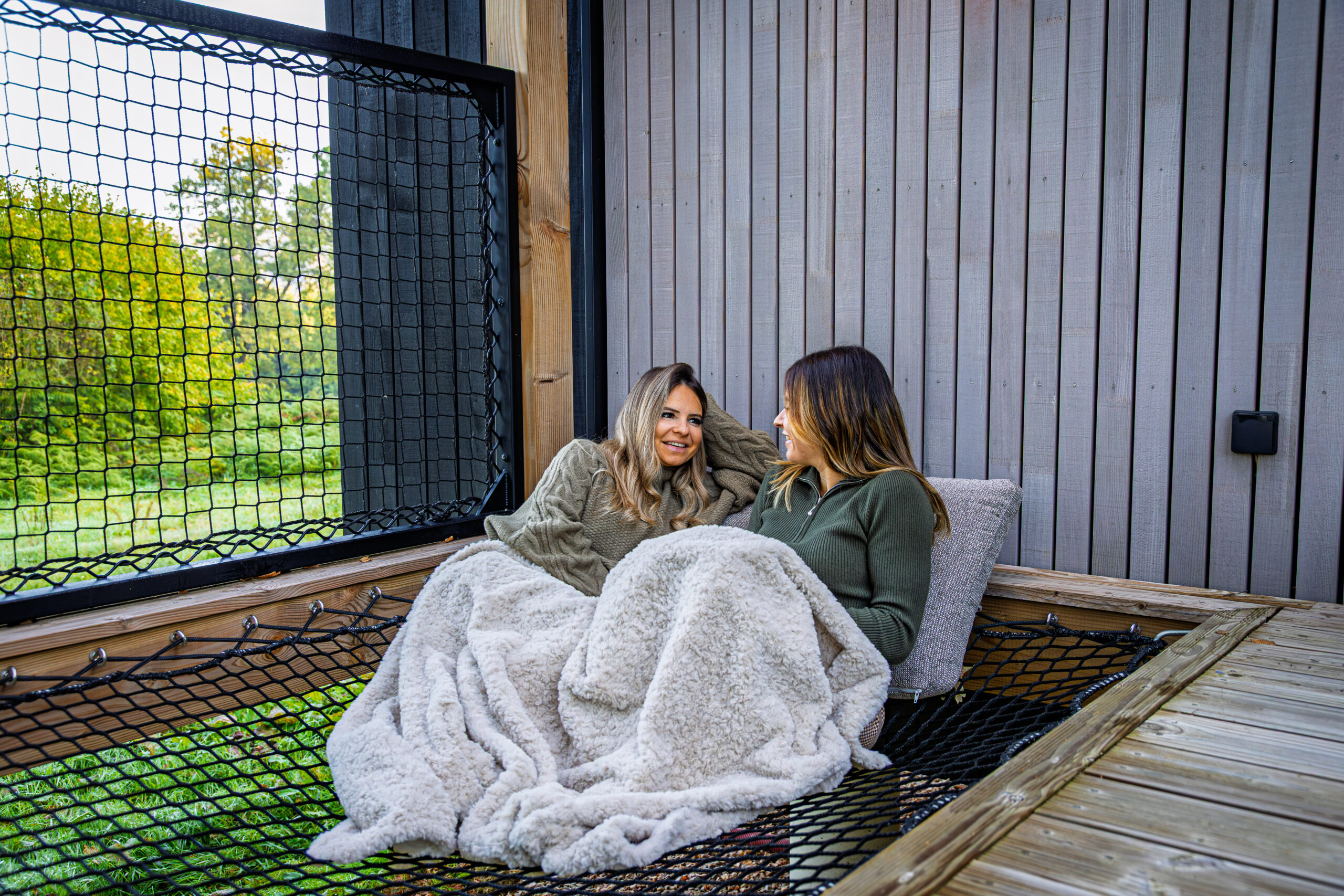 Deux femmes détendues sous un plaid sur le filet suspendu de la terrasse d'un lodge Les Hauts du Val, vue sur la verdure