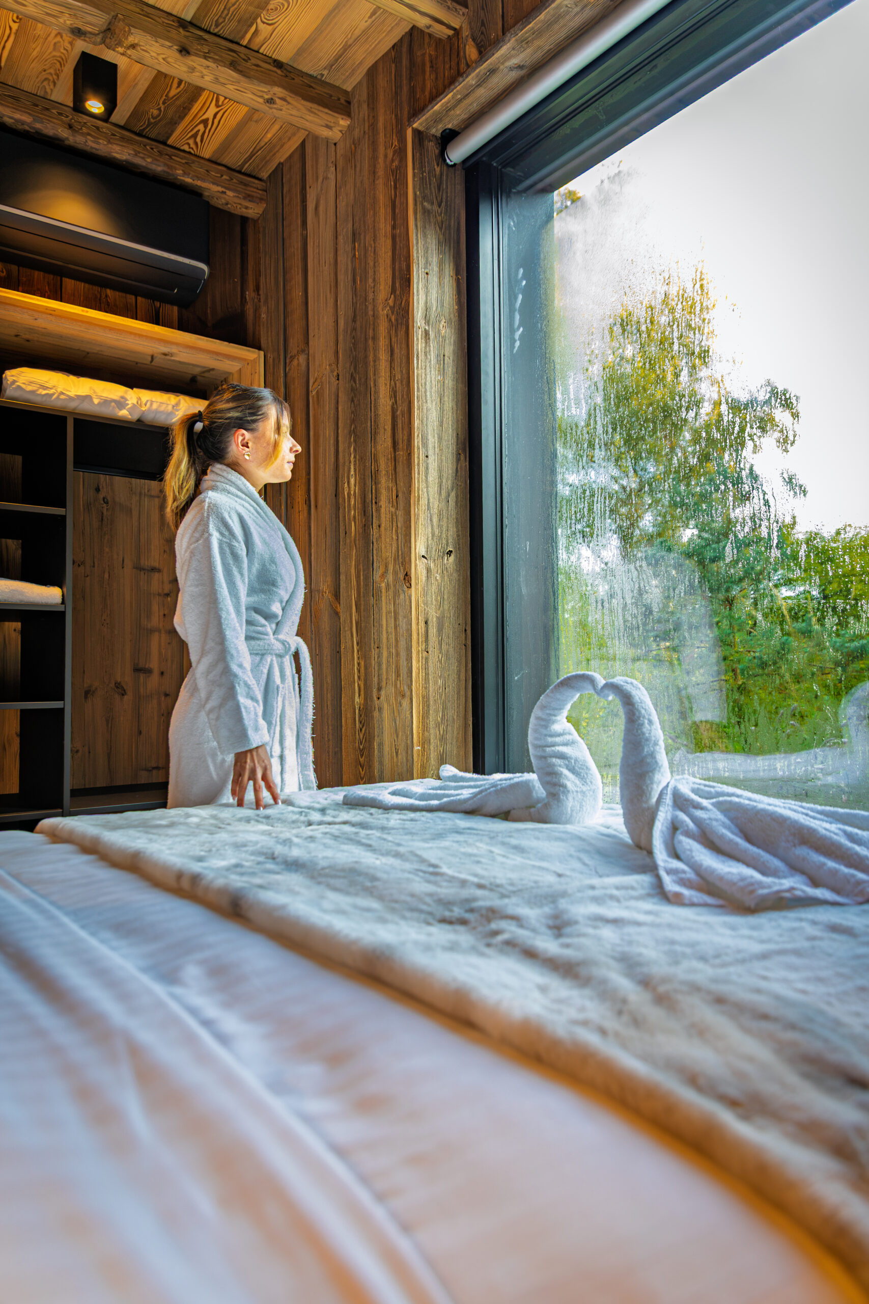 Femme en peignoir devant la grande baie vitrée d'une chambre de lodge Les Hauts du Val, serviettes pliées en cygnes sur le lit